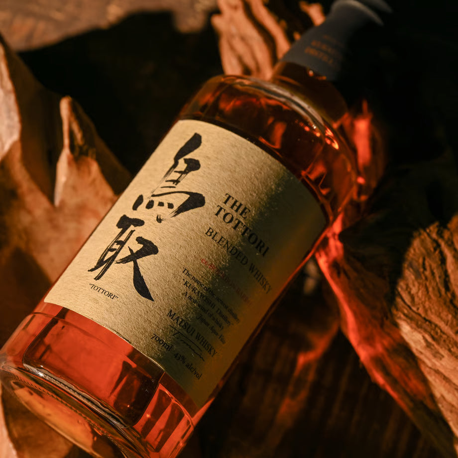 マツイブレンデッドウイスキー「鳥取 金ラベル」のボトルが木の上に横たわる写真。モルトとグレーン原酒を合わせ、バーボン樽由来の甘い香りが特徴のウイスキー。