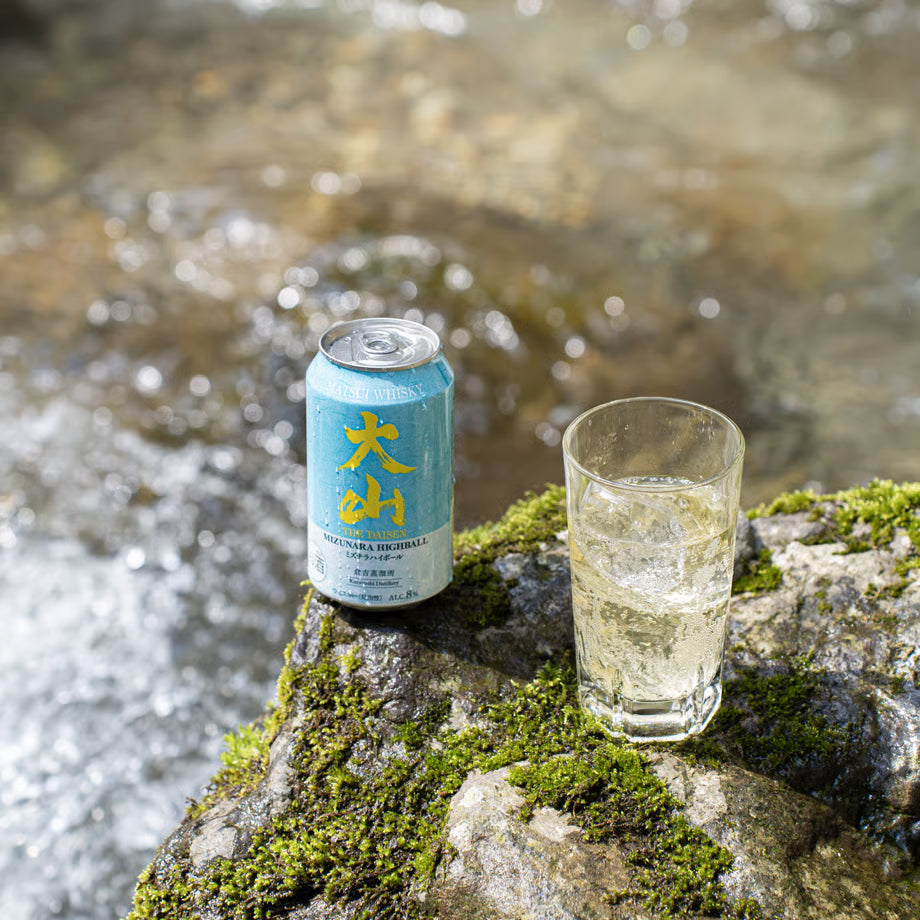 大山ミズナラハイボールの缶とグラスを、清流のそばの岩場に置いたイメージカット。自然の中で楽しむ爽やかな味わいを表現した350ml・8%のハイボール。
