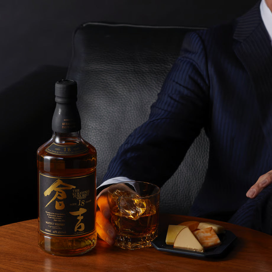 マツイピュアモルトウイスキー「倉吉18年」とロックグラス、チーズの盛り合わせがテーブルに置かれ、男性がグラスに手を添えている写真。落ち着いた雰囲気の一枚。